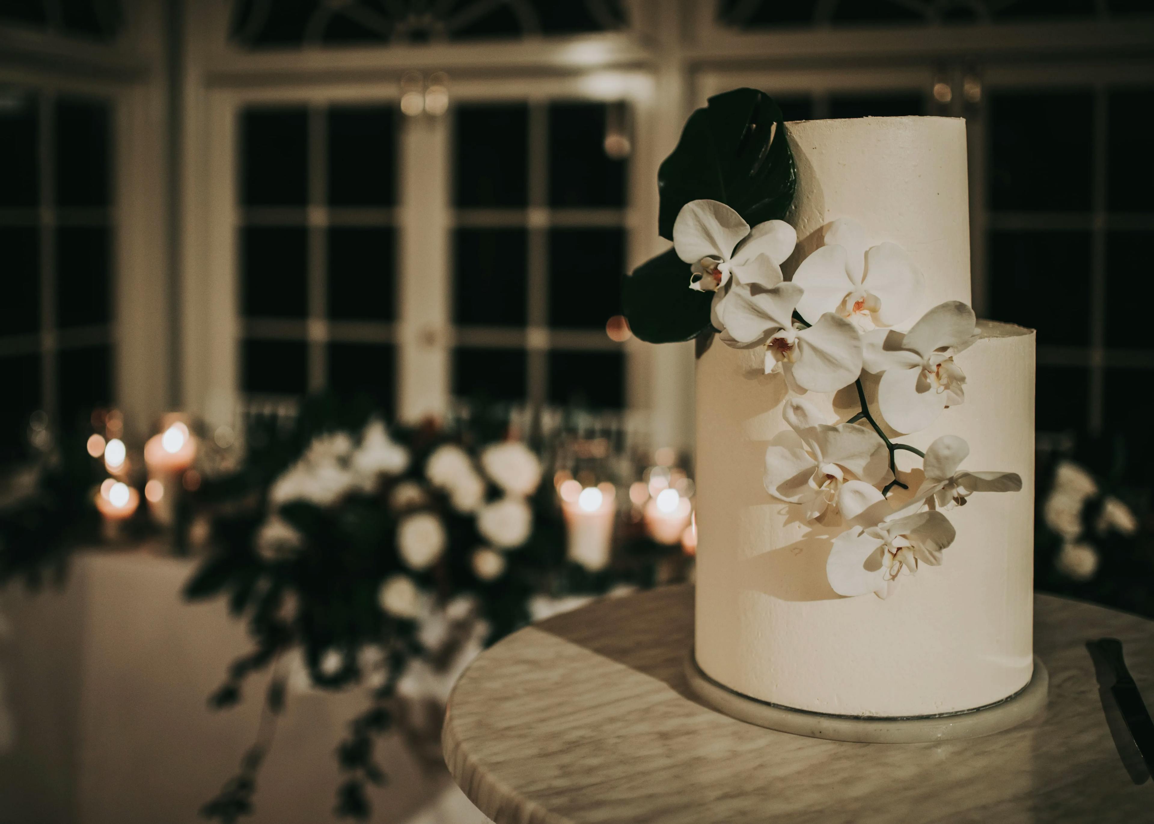 A white, two-tiered cake adorned with white flowers and green leaves sits on a round table. The cake is in front of a window with curved panes, and the background features a dimly-lit room with candles and floral arrangements.