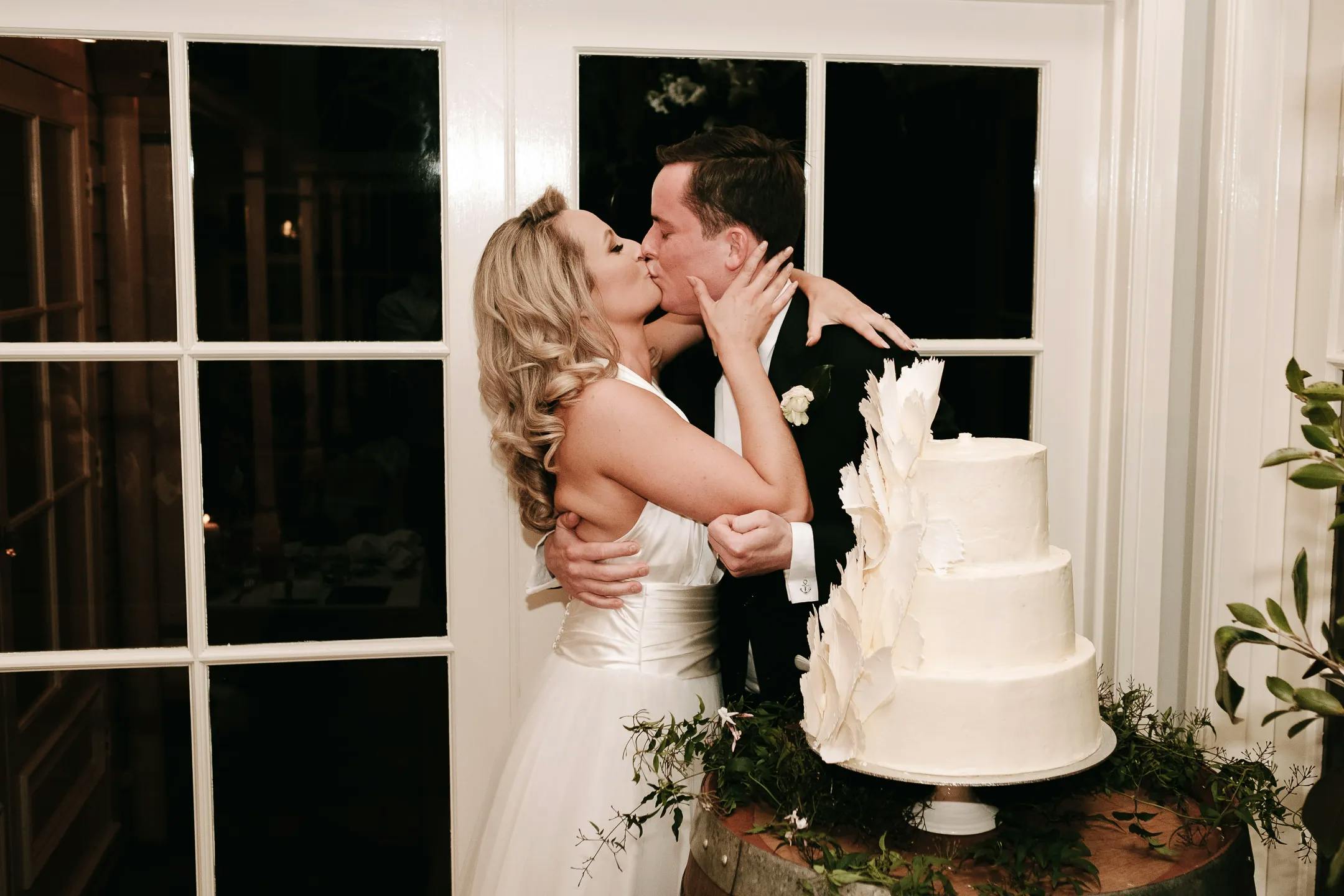 A newlywed couple shares a kiss while embracing next to a three-tiered white wedding cake adorned with leaf-like decorations. They are standing in front of large glass windows in a warmly lit room. The bride is in a strapless white dress, and the groom is in a dark suit.