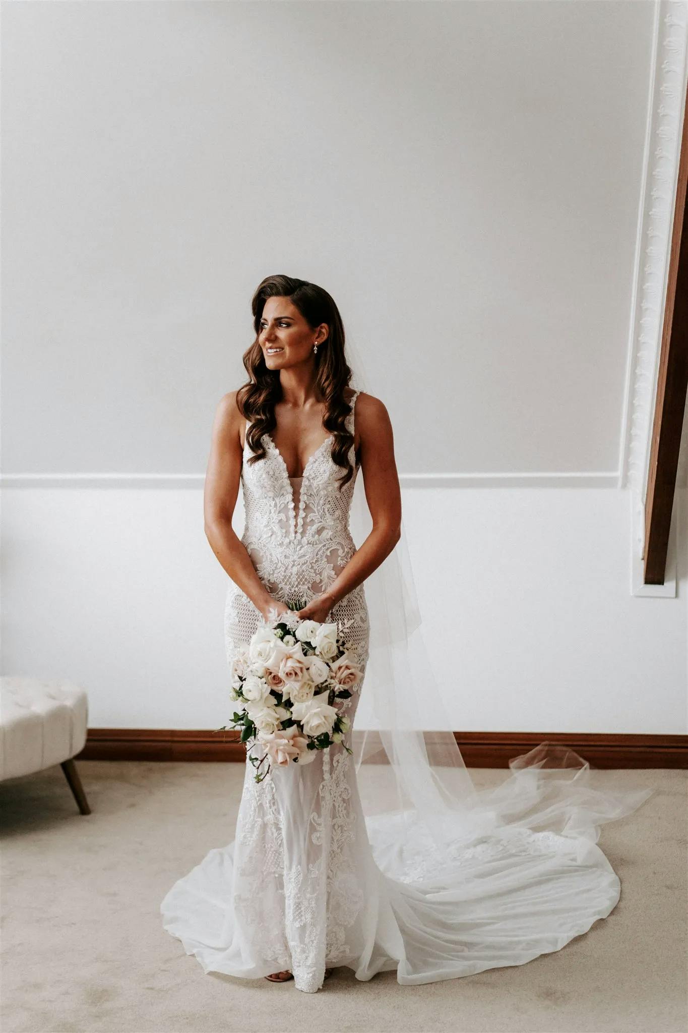 A bride in a white lace wedding dress and veil, holding a bouquet of white and blush flowers. She is standing indoors against a minimalist background and smiling slightly to the side. The train of her dress is spread out behind her on the floor.