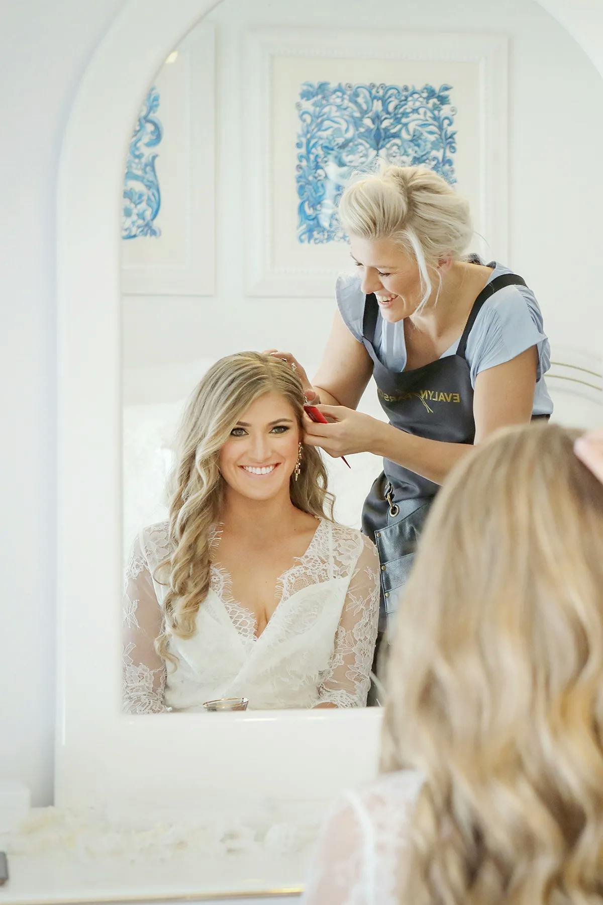 A smiling bride with long, wavy blonde hair and a lace bridal gown sits in front of a mirror. A hairstylist stands behind her, styling her hair with a curling iron. The reflection captures the happy moment in a bright, white room with elegant decorations.