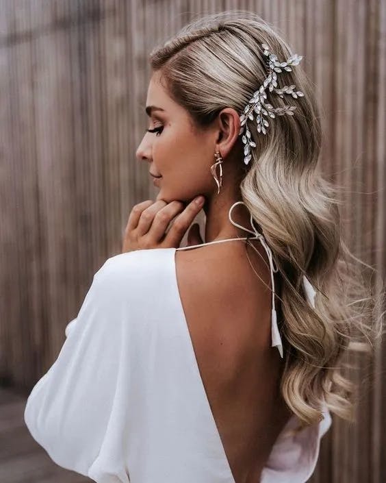 A woman with long, wavy blonde hair is shown from behind, with her head turned slightly to the side. She is wearing a white, open-back dress and a delicate hair accessory adorned with crystals. She has a pair of dangling earrings and is posing with one hand near her face.