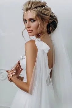 A bride with her hair styled in an elegant updo, adorned with a delicate headpiece, poses in her white wedding dress. The sheer veil cascades down her back, and her expression is serene as she glances over her shoulder. The background is softly lit and neutral.