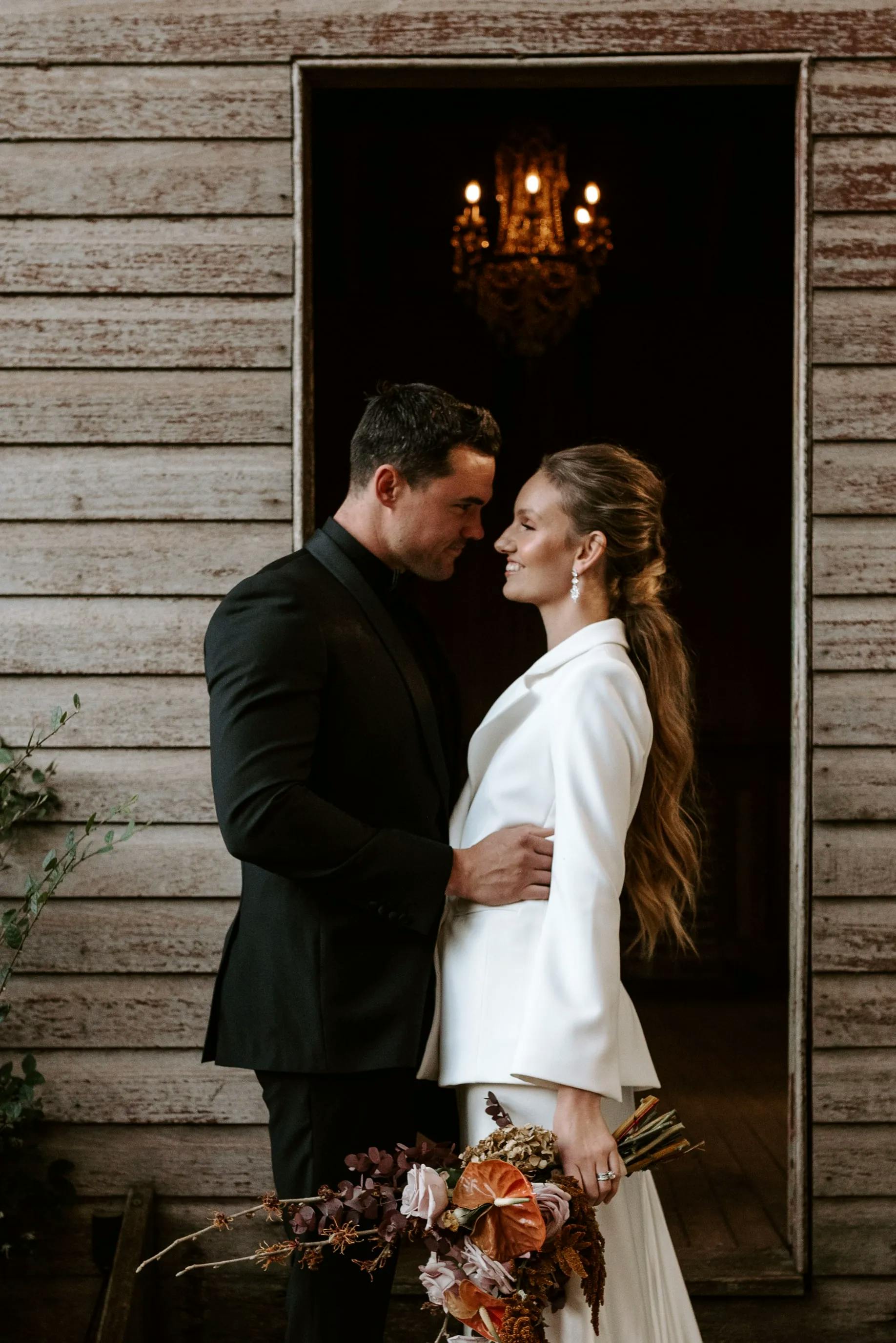 A couple embraces in front of a rustic wooden wall with a dark open doorway behind them. The man is dressed in a black suit, while the woman wears a white outfit and holds a bouquet of flowers. A chandelier is visible in the background.