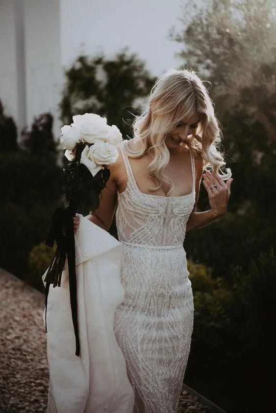 A bride in a lacy, fitted wedding dress with thin straps holds a bouquet of white roses and a long, white veil over her arm. Her blonde hair cascades in soft waves as she looks down, walking on a gravel path with greenery in the background.