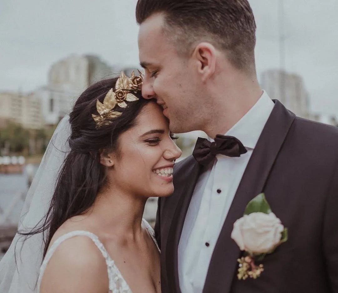 A bride and groom share an intimate moment outdoors. The groom, in a dark suit with a bow tie, gently kisses the bride on the forehead. The smiling bride wears a white dress, veil, and floral headpiece. Urban buildings are visible in the background.