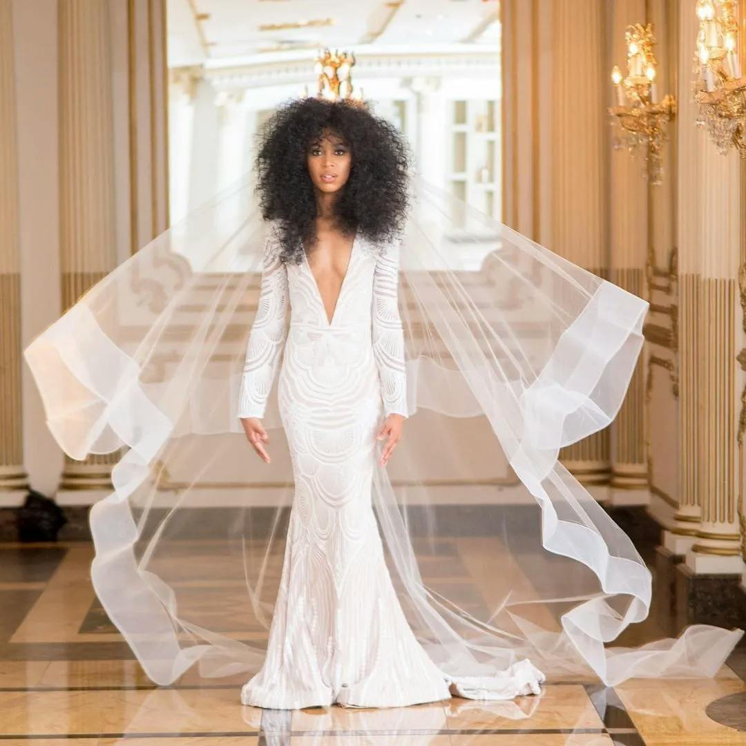 A bride in a luxurious setting, wearing a fitted, long-sleeve white wedding dress with an intricate pattern and a deep V-neckline. She has curly hair and is adorned with a flowing veil, standing on a polished marble floor with opulent, gold-accented decor.