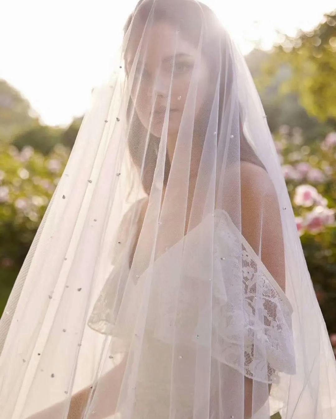 A bride stands outdoors on a sunny day, wearing a white lace wedding dress and a long, sheer veil with small embellishments. Soft sunlight filters through the veil, illuminating her face. The background features green foliage and blooming pink flowers.