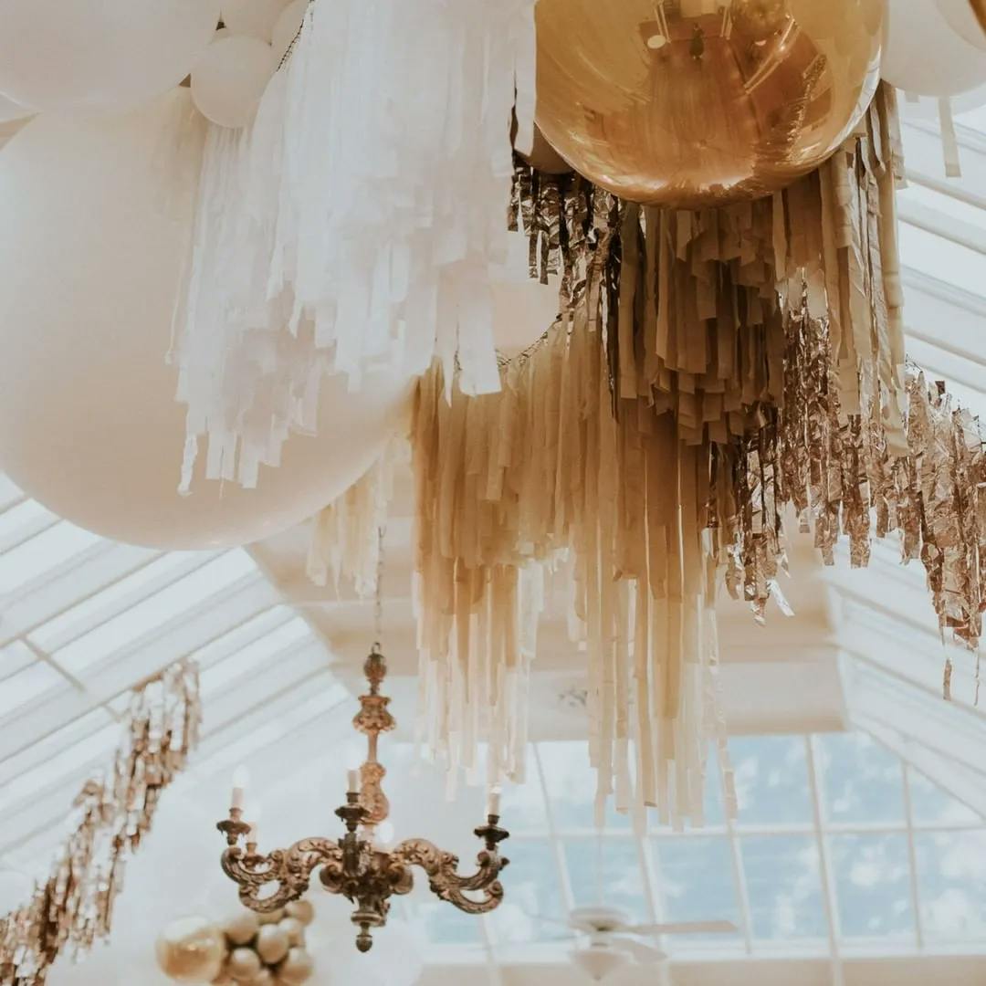 An elegant ceiling display features large white and gold balloons along with white, gold, and silver fringe streamers. An ornate chandelier hangs alongside the decorations in a room with a high, glass-paneled ceiling.