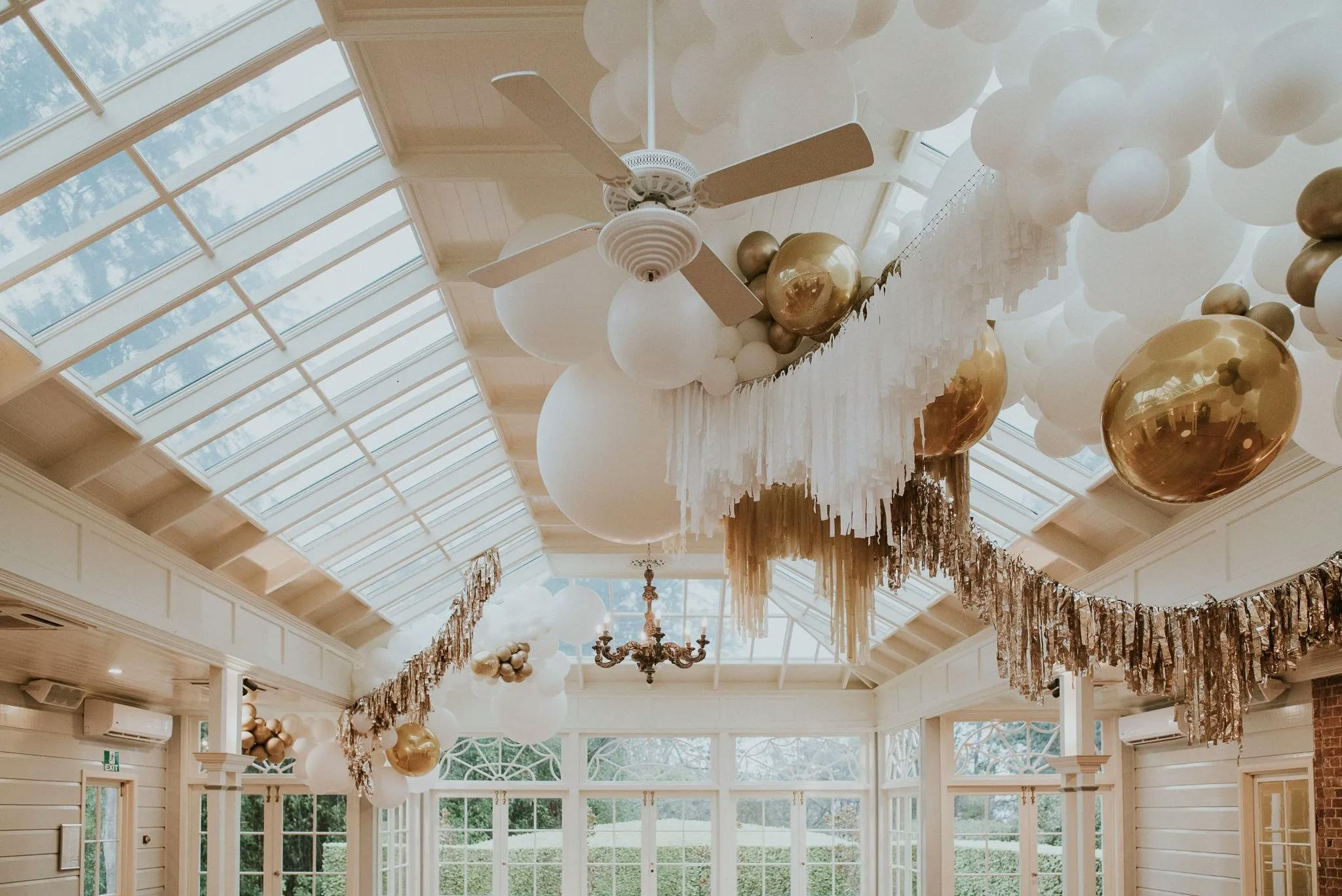 An elegant room with a high glass ceiling decorated with large white and gold balloons, white tassels, and gold garlands. The room is bright and airy, with chandeliers hanging from the ceiling, and large windows that let in natural light.