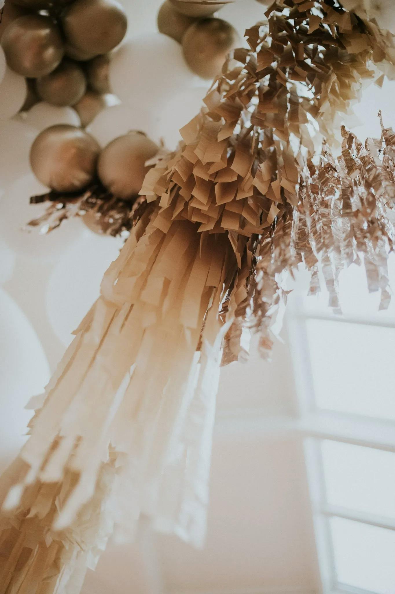 Close-up view of a decorative hanging installation featuring cascading beige and golden fringed paper. In the background, several inflated gold balloons can be seen against a softly lit, out-of-focus backdrop. The setup evokes a festive and elegant ambiance.