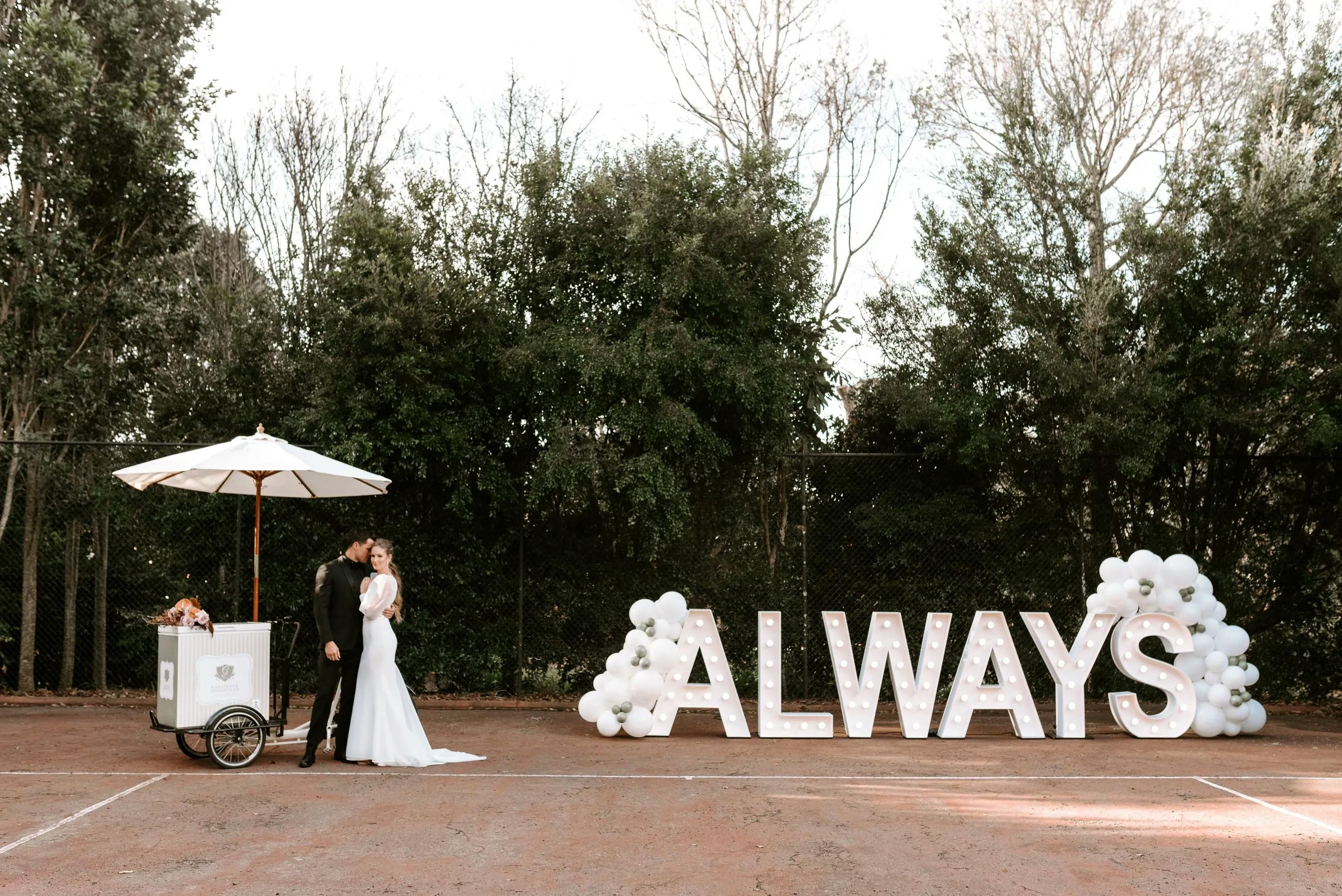 A bride and groom share a kiss next to a decorated ice cream cart with a white umbrella. Large illuminated letters spell out "ALWAYS," decorated with white balloons and flowers, set against a backdrop of trees.