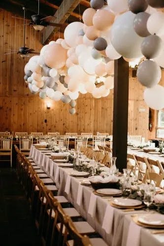 A long banquet table is decorated with white tablecloths, elegant tableware, and floral centerpieces. Overhead, a large arrangement of white, gray, and blush pink balloons hangs from the wooden ceiling. Wooden walls and chairs create a rustic ambiance.