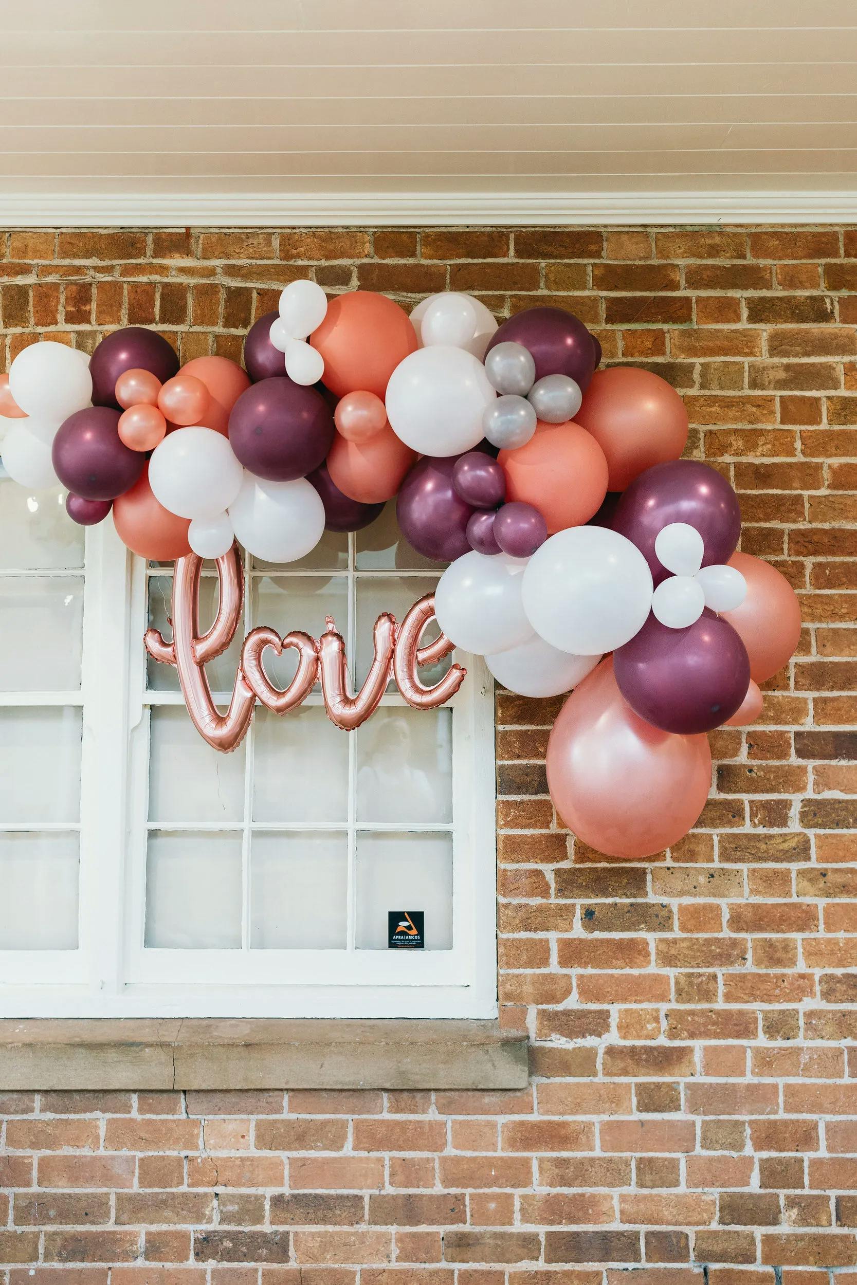 A decorative arrangement of white, pink, peach, and purple balloons is arching over a window against a brick wall. In the middle of the balloon arch hangs a shiny metallic pink balloon spelling "love" with a heart shape forming the letter O.