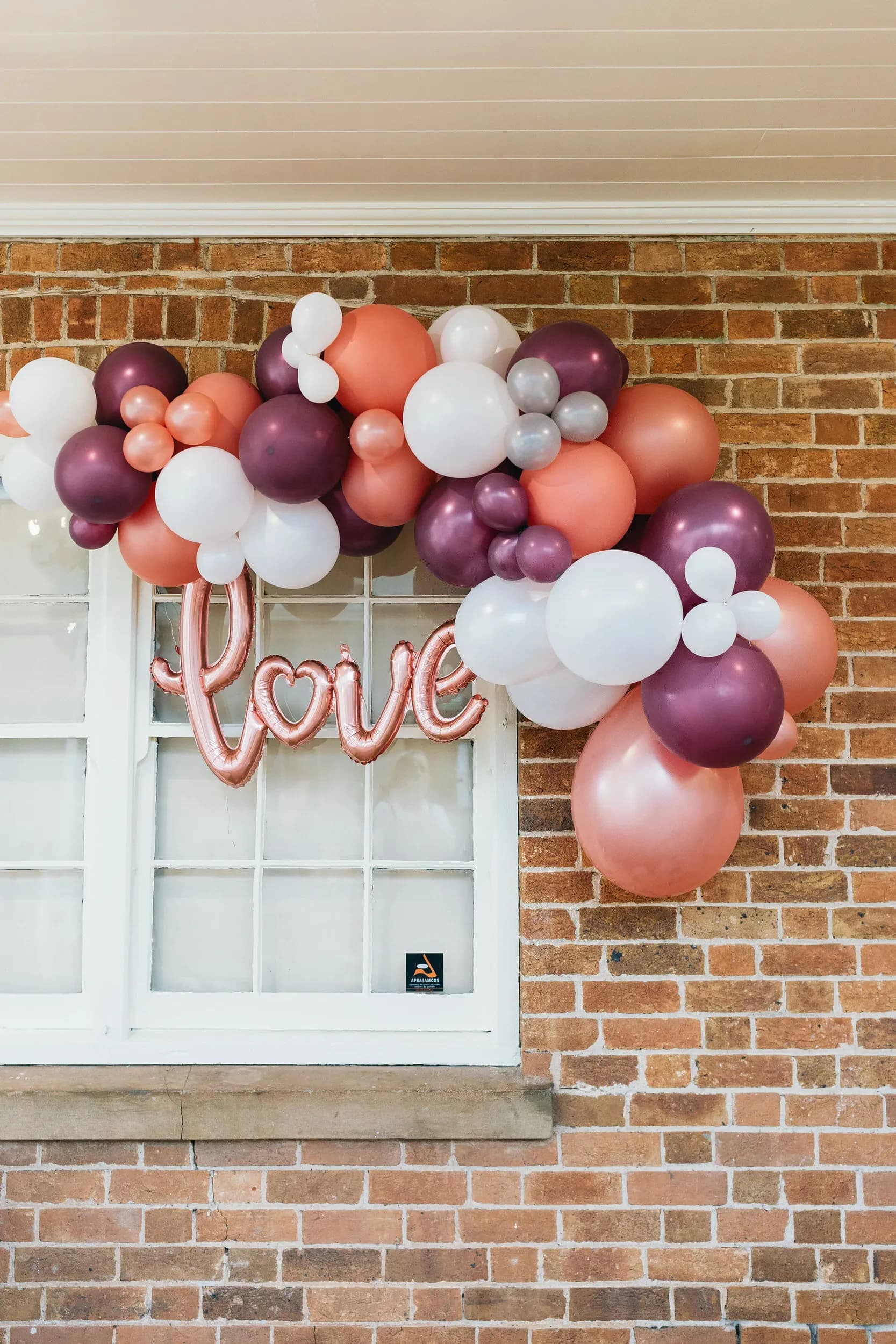 A decorative arrangement of white, pink, peach, and purple balloons is arching over a window against a brick wall. In the middle of the balloon arch hangs a shiny metallic pink balloon spelling "love" with a heart shape forming the letter O.