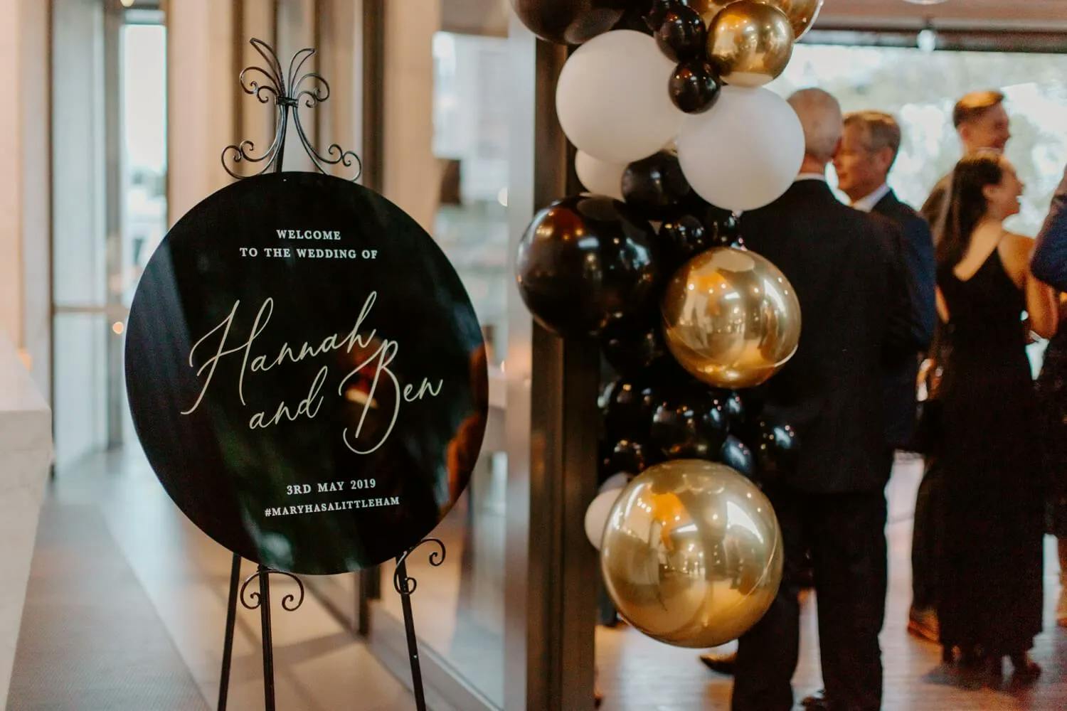 An elegant wedding display featuring a black circular welcome sign with white cursive text that reads "Welcome to the wedding of Hannah and Ben" and a date. Black, white, and gold balloons are arranged next to the sign. Guests in formal attire mill around in the background.