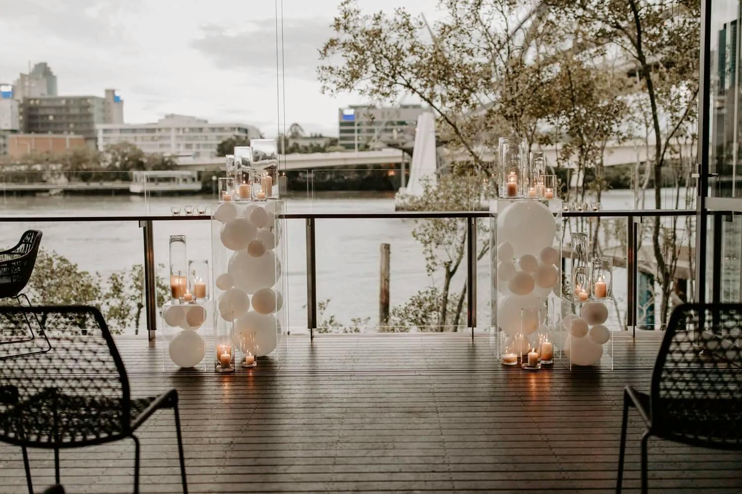 A serene riverside setting features a wooden deck adorned with white balloon columns and numerous candles. Clear glass vases brimming with candles line the deck. Modern black wicker chairs are neatly arranged, while city buildings and leafless trees frame the background.