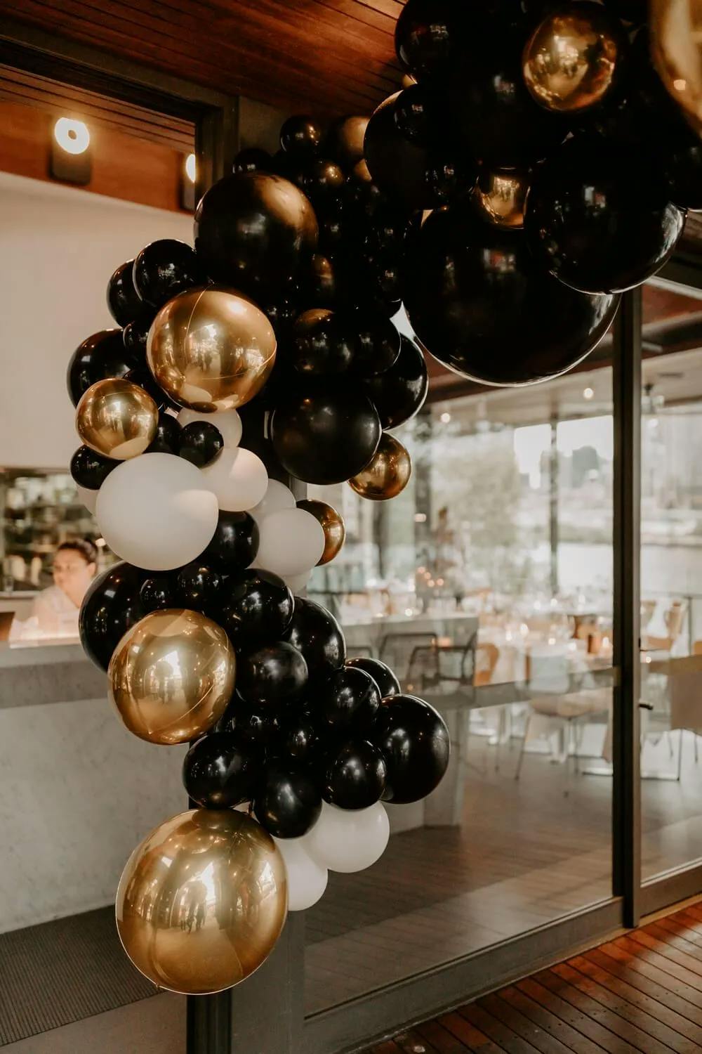 A decorative arch made of black, gold, and white balloons is displayed near the entrance of a modern indoor event space. The interior features glass walls, wooden accents, and a view of dining tables set up with elegant place settings.