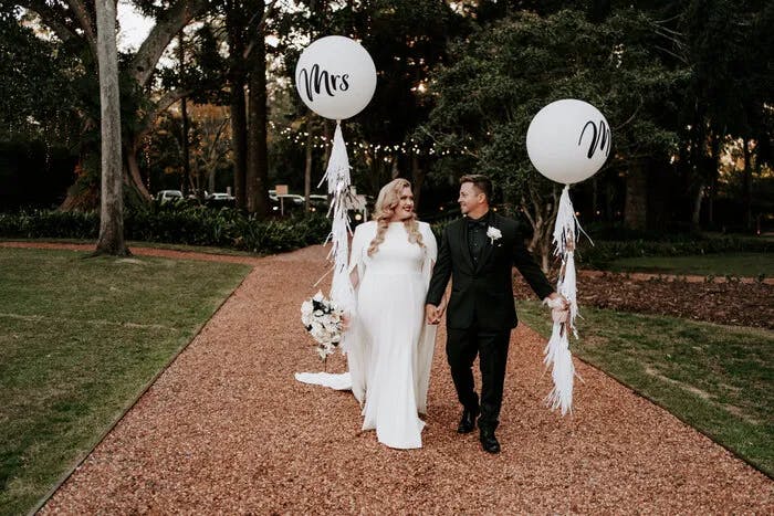 A bride and groom walk hand in hand on a gravel path in a garden. The bride, in a white dress, holds a balloon labeled "Mrs," and the groom, in a black suit, holds a balloon labeled "Mr." They both smile at each other while surrounded by trees and greenery.