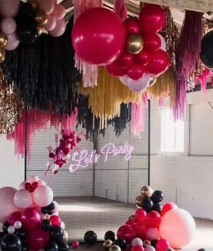A party setup with colorful balloon arrangements, predominantly in pink, black, and gold. A neon sign reading "Let's Party" is illuminated in the middle. The ceiling is decorated with streamers and the scene is well-lit with natural light from a window.