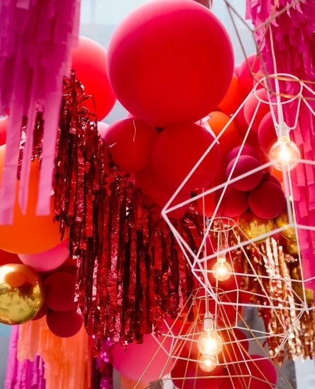 A vibrant and festive scene with large red and pink balloons, metallic red fringe garlands, and geometric light fixtures with exposed bulbs. Pink fringed streamers hang from above, adding to the lively and colorful atmosphere.