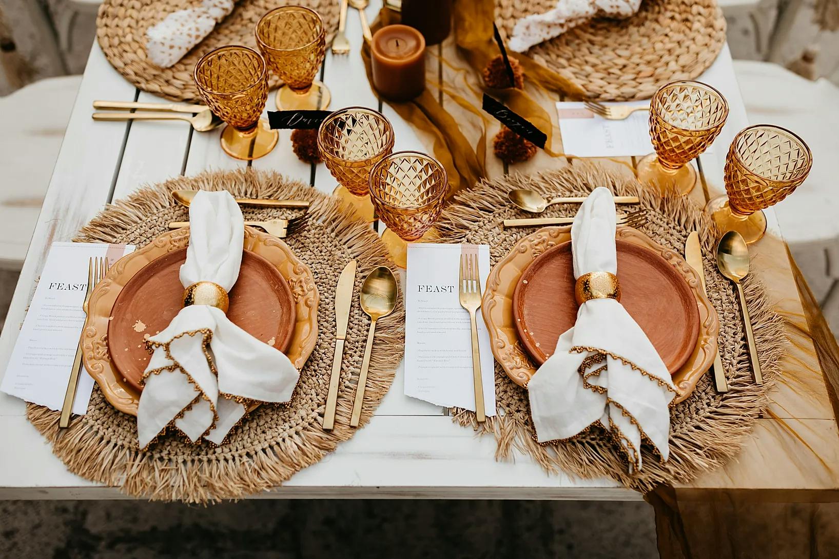 A stylishly set table for two, featuring woven placemats, terra-cotta plates, gold utensils, amber glasses, and white napkins with gold napkin rings. There are menus labeled "FEAST" and a small candle in the center, enhancing the elegant dining setup.