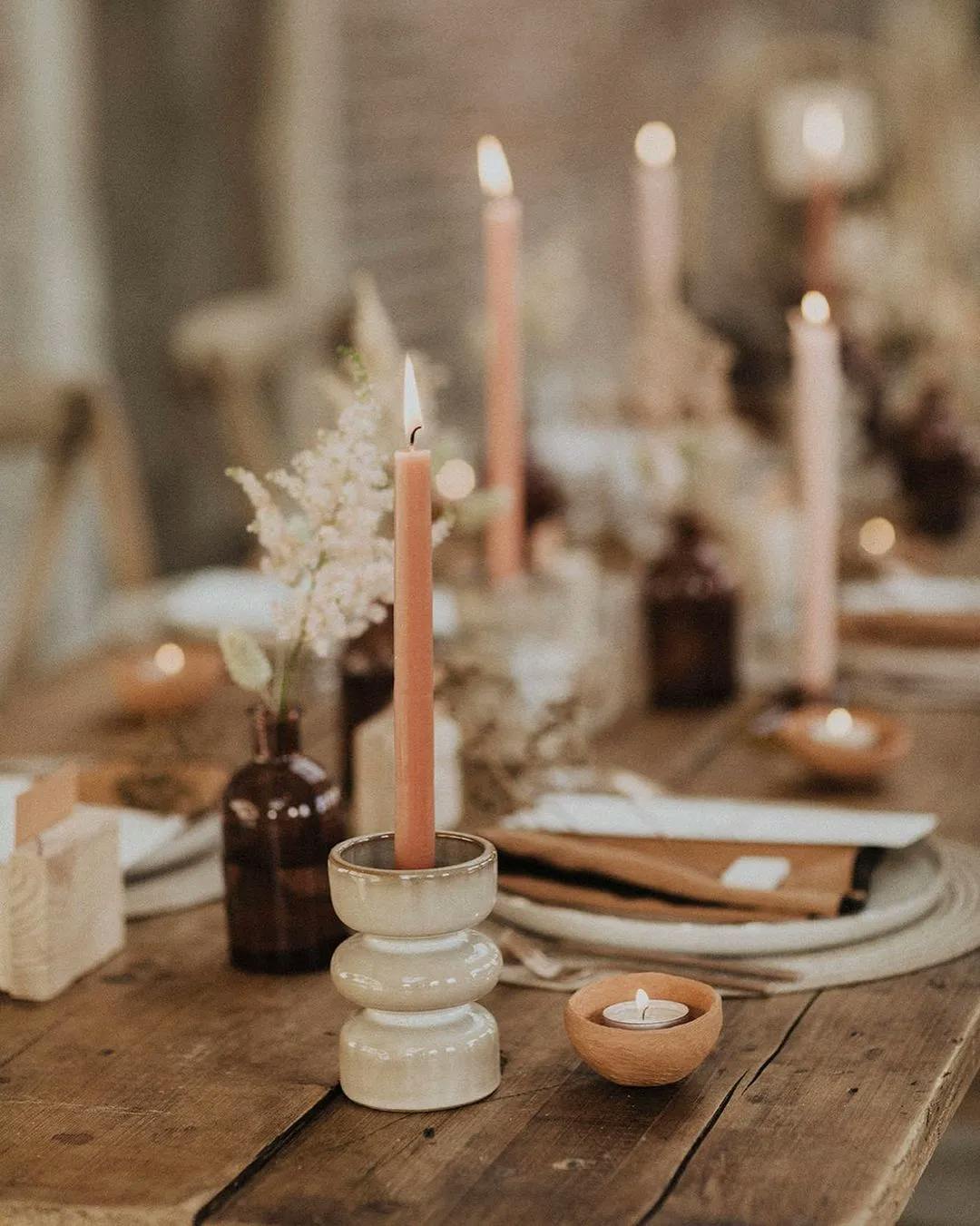 A rustic wooden table is adorned with lit pink candles in ceramic holders, small wooden bowls with tea lights, and delicate floral arrangements in small brown vases. The table setting includes earth-toned napkins and plates, creating a warm and inviting atmosphere.