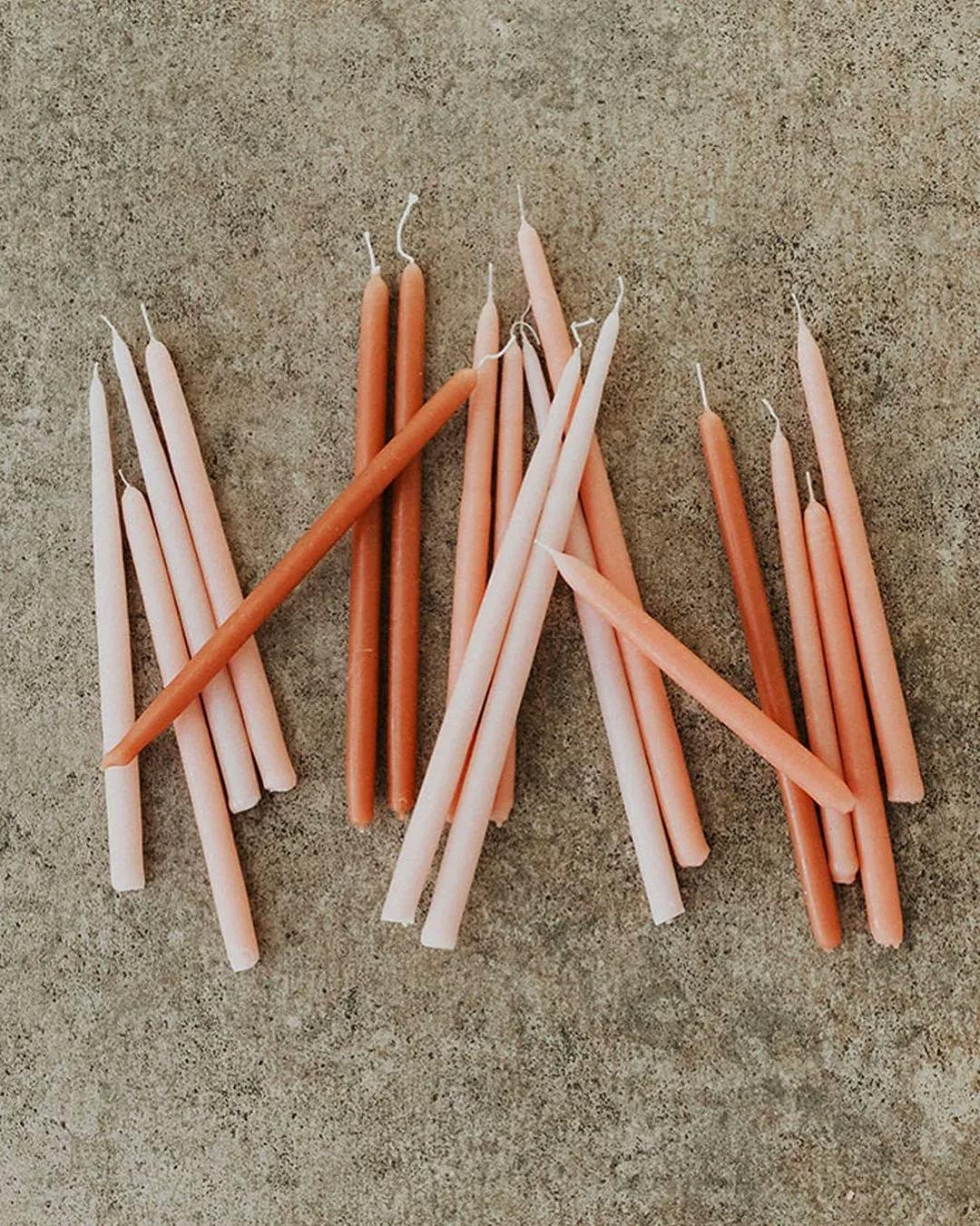 A collection of tapered candles in various shades of pink and orange are scattered on a textured, light brown surface. The candles vary slightly in length and thickness, and some have visible wicks while others do not.