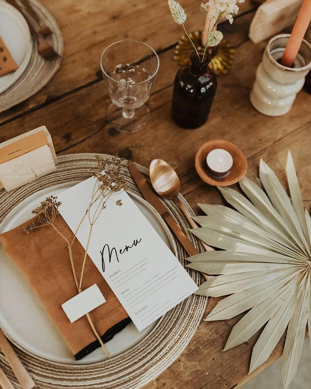 A rustic table setting features a white plate with a menu, a folded brown napkin, and dried botanical accents. Nearby are a clear glass, brown bottle with dried flowers, candleholder, and a dried palm leaf on the right side of the wooden table.