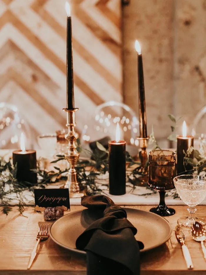 A stylishly set dining table with black candles in gold holders, a black napkin on a tan plate, and elegant glassware. The table is adorned with green foliage and small decor pieces, creating a cozy, intimate ambiance.