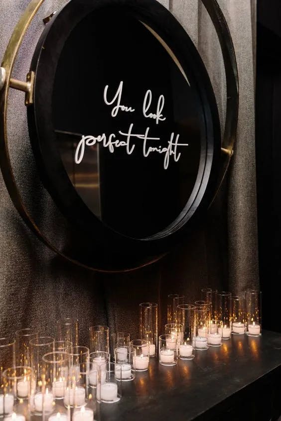 A glowing neon sign that reads "You look perfect tonight" is displayed on a circular mirror with a metal frame. Below the sign, numerous small candles in glass holders are arranged, casting a warm and cozy ambiance.