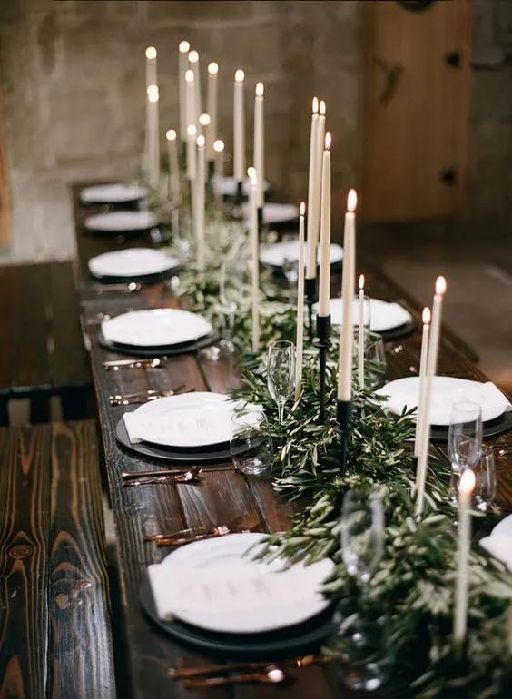 A rustic wooden dining table is elegantly set for a formal event. Tall, white taper candles in black holders and green foliage form a centerpiece. Each place setting includes white plates, silver cutlery, and glassware, under soft, ambient lighting.