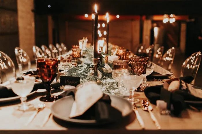 A long, elegantly set dinner table with black candles, decorative greenery, and an array of crystal glasses and cutlery. Warm lighting enhances the ambiance, with gold and black accents creating a sophisticated atmosphere, perfect for a formal gathering.