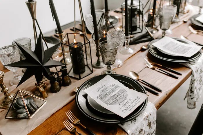 A rustic wooden dining table is elegantly set with black and gold dinnerware, black candles in geometric candle holders, and glass goblets. Each place setting includes a handwritten menu. Decorative black and gold ornaments add to the sophisticated ambiance.