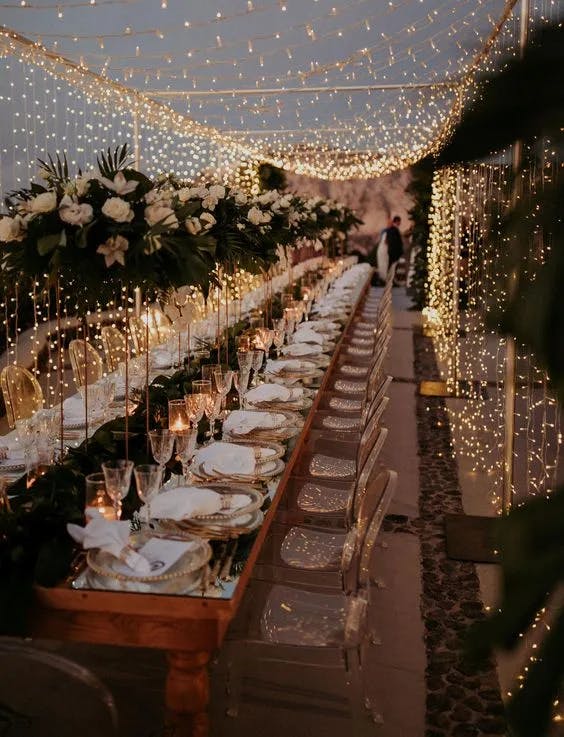 A long, elegant, outdoor wedding reception table is adorned with white flowers and greenery, set under a canopy of twinkling string lights. Delicate table settings include clear glassware and white plates. Two people are visible in the background, blurred.