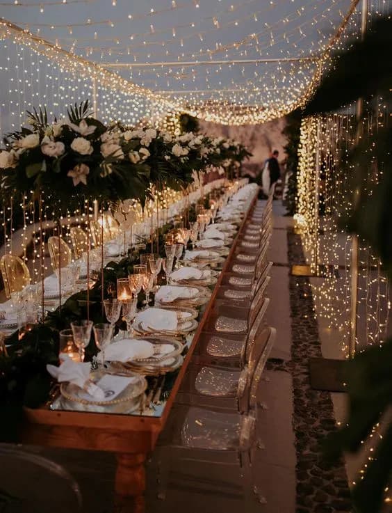 A long, elegant, outdoor wedding reception table is adorned with white flowers and greenery, set under a canopy of twinkling string lights. Delicate table settings include clear glassware and white plates. Two people are visible in the background, blurred.
