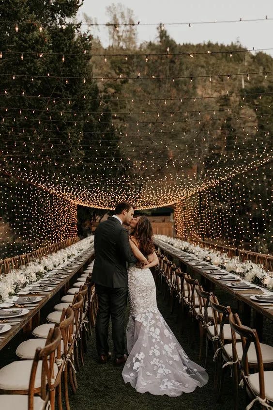 A couple stands embraced at the center of a beautifully decorated outdoor reception with long tables arranged symmetrically. String lights hang above, creating a canopy effect. The bride's white wedding dress contrasts with the groom's dark suit. Trees border the area.