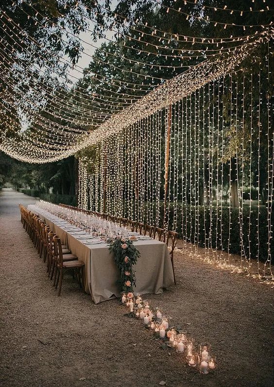 A long outdoor dining table is beautifully decorated with a beige tablecloth, wooden chairs, and a floral centerpiece at one end. Overhead and down the sides, warm string lights create a magical, intimate ambiance. Candles in glass holders line the path beside the table.