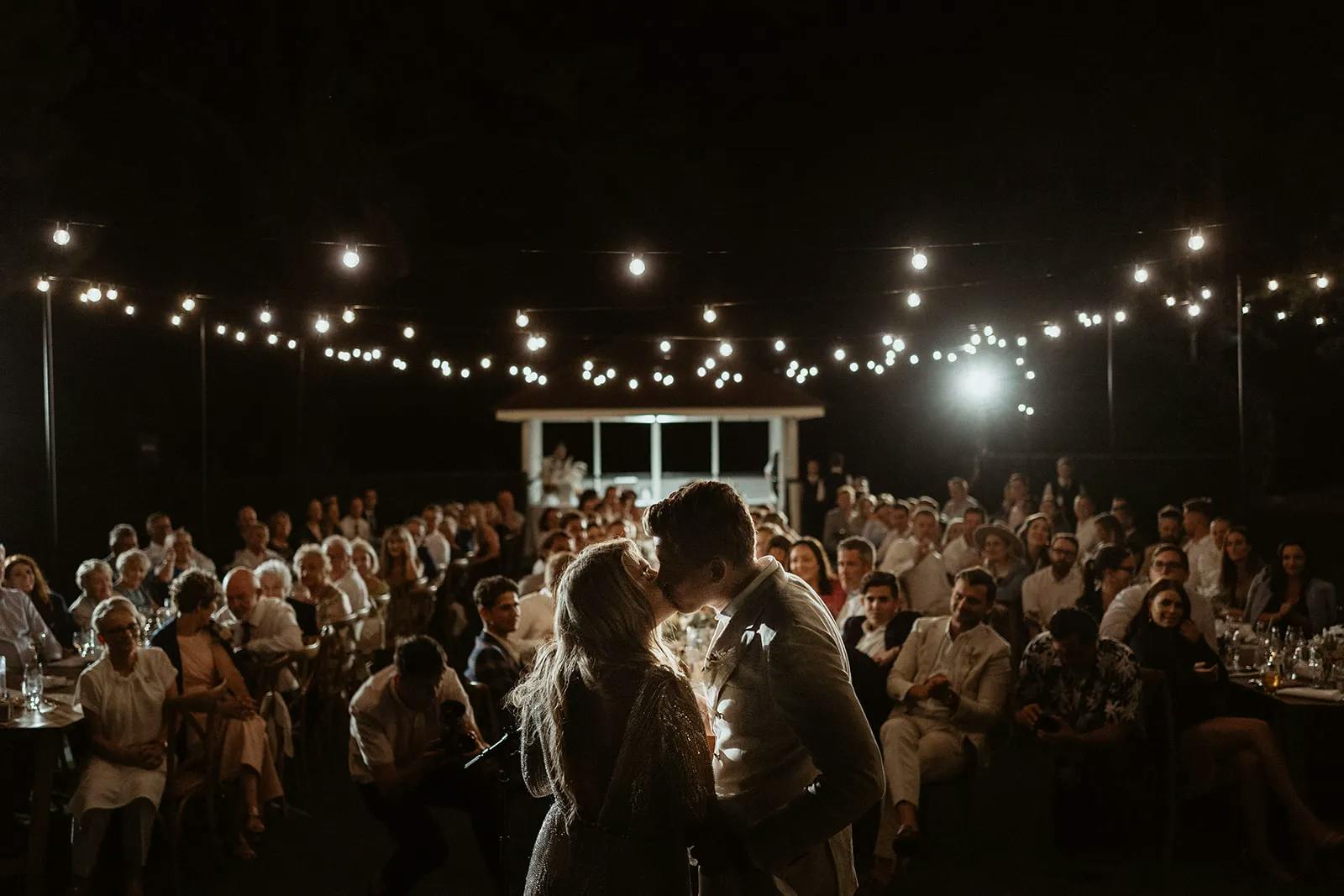 A couple shares a kiss under string lights, surrounded by a large seated audience in an outdoor nighttime setting. The scene has a warm, intimate atmosphere, with fairy lights casting a gentle glow over the gathering.