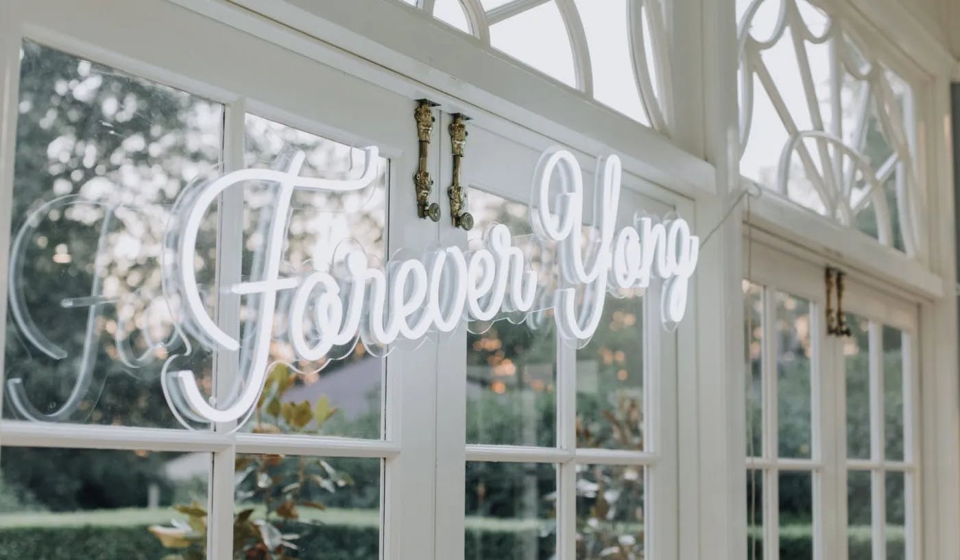 Neon sign reading "Forever Young" mounted on large windows inside a bright, elegant room with ornate detailing. The view outside the window shows greenery and a soft, evening glow.
