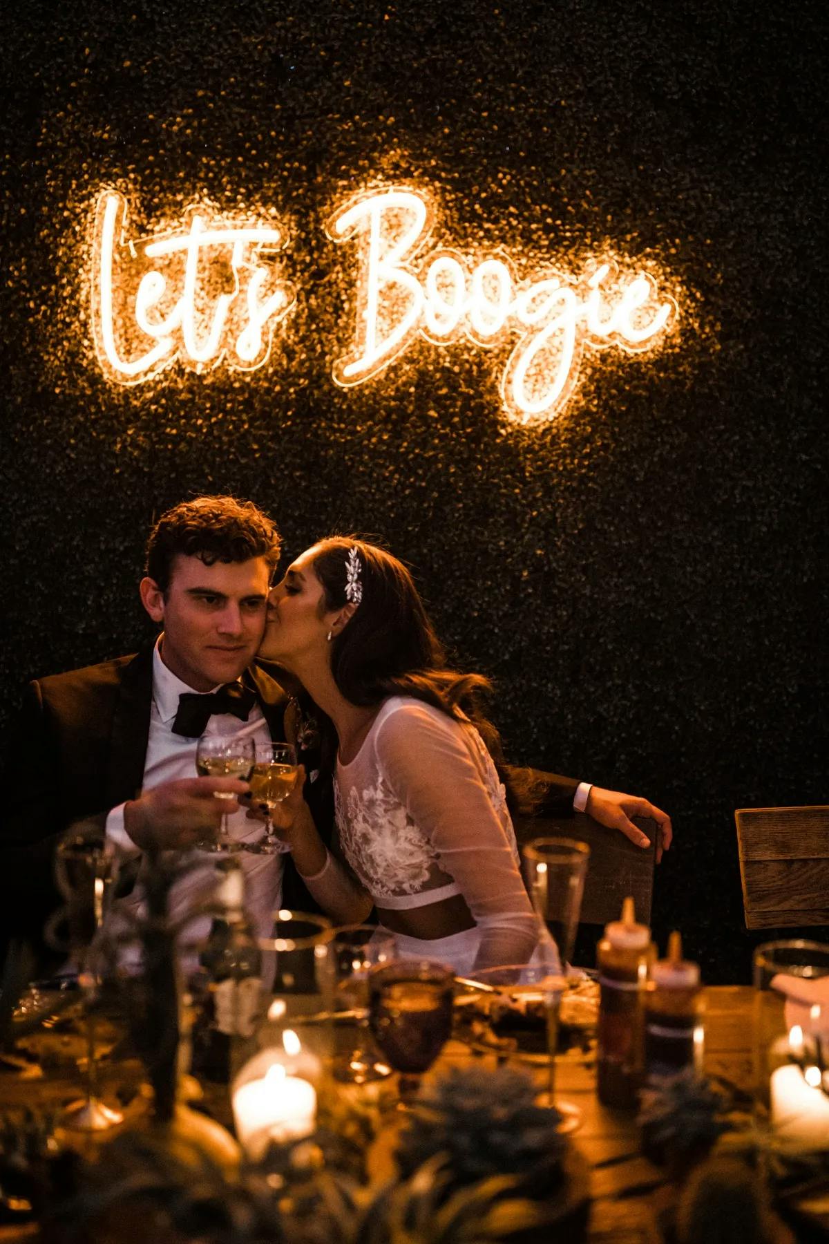 A couple sits closely at a dimly lit wedding reception. The woman kisses the man's cheek while he holds a glass. Behind them, a neon sign reads "Let's Boogie." The table is set with wine glasses, condiments, and decor. The ambiance is warm and festive.