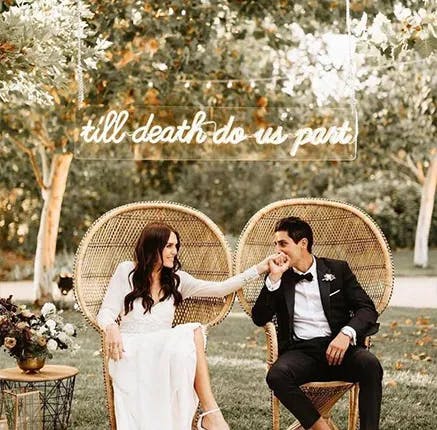 A bride and groom are seated on elegant wicker chairs outdoors under trees. The bride is dressed in a white wedding gown, while the groom wears a black tuxedo. They appear happy, holding hands and smiling, with a neon sign above them reading "till death do us part.