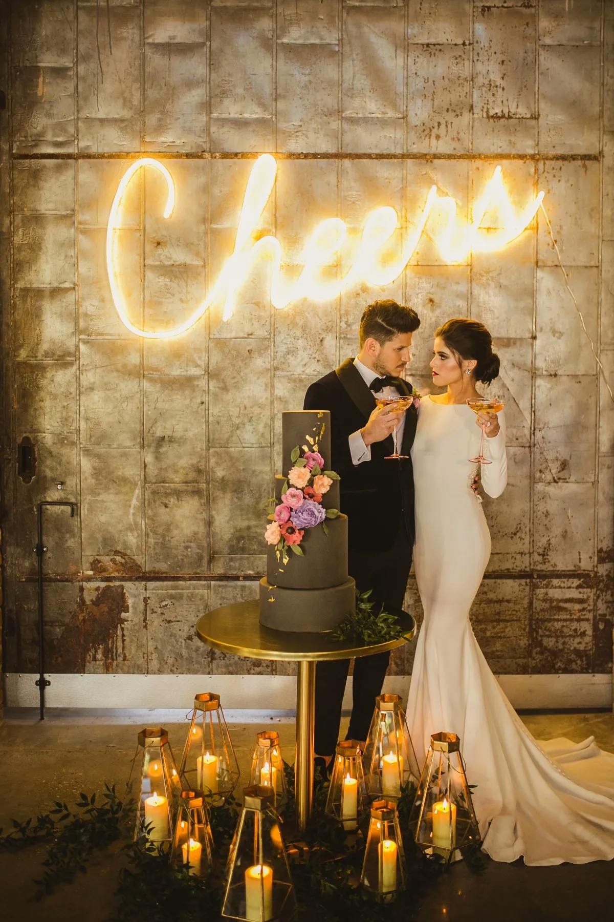 A bride and groom in formal attire stand close together, holding champagne glasses in a warmly lit room. They are in front of a dark wedding cake adorned with flowers, and a bright neon sign that reads "Cheers" is on the wall behind them. Lanterns with candles surround them.