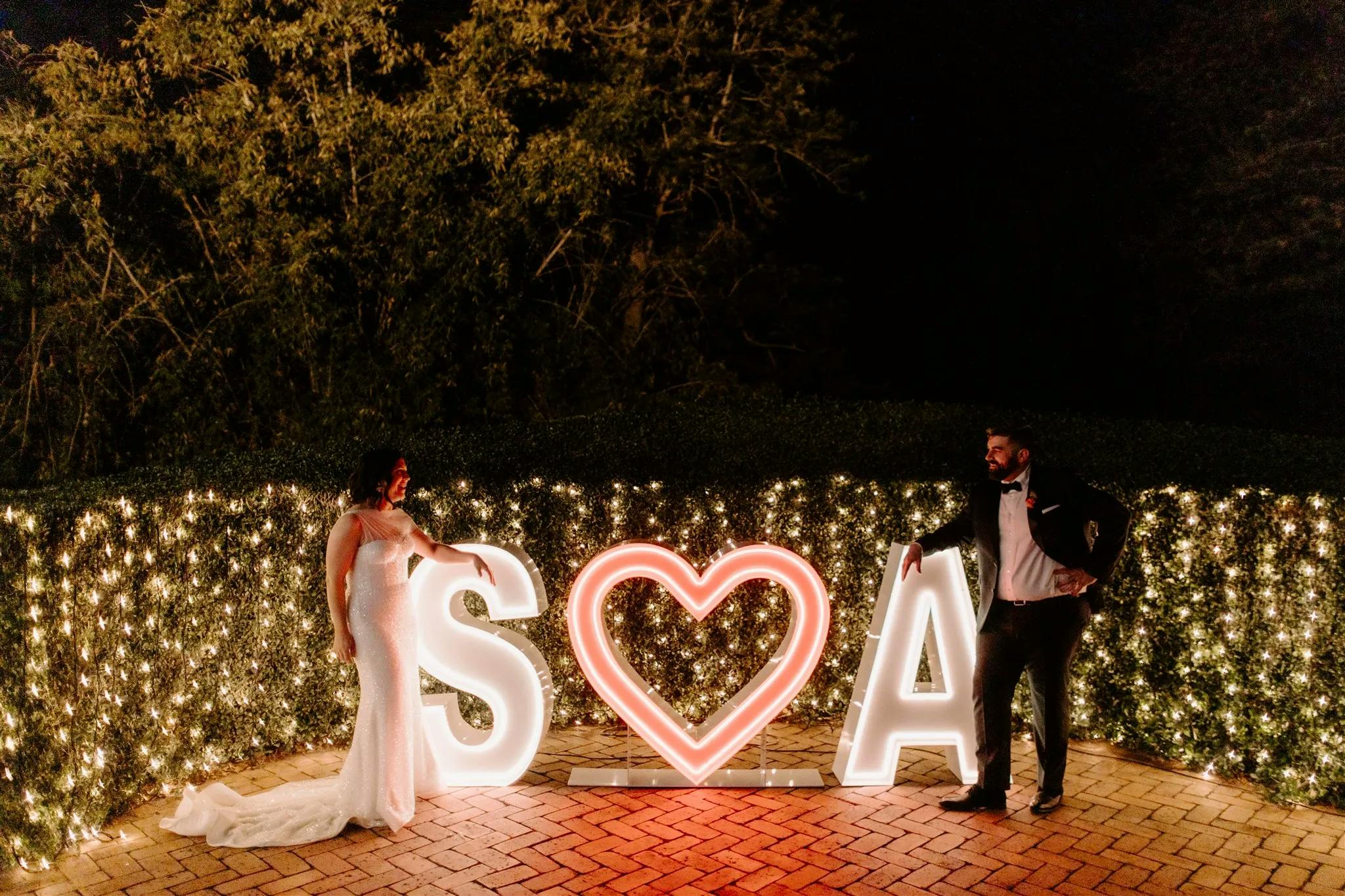 A couple, dressed in formal wedding attire, stands on either side of illuminated letters spelling "S (heart) A" in front of a hedge adorned with fairy lights. The bride is on the left in a white dress, and the groom is on the right wearing a tuxedo.