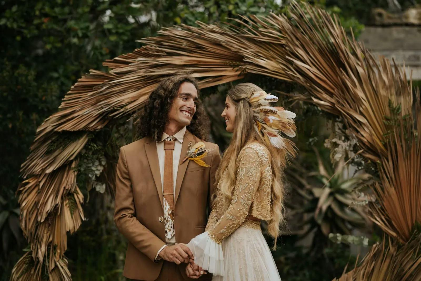 A couple stands holding hands and smiling at each other in front of a rustic, circular arbor made of dried palm leaves. The man wears a brown suit with a floral tie and boutonnière, while the woman dons a lace top, tulle skirt, and feather hair accessories.