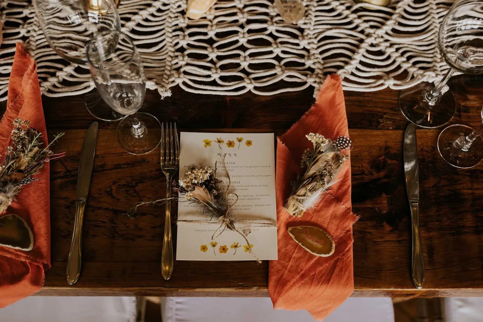 A rustic wooden dining table set for an event with elegant tableware. The setup includes a menu card decorated with dried flowers, gold cutlery, orange napkins, glasses, and small decorative items. A macrame table runner adds a bohemian touch.