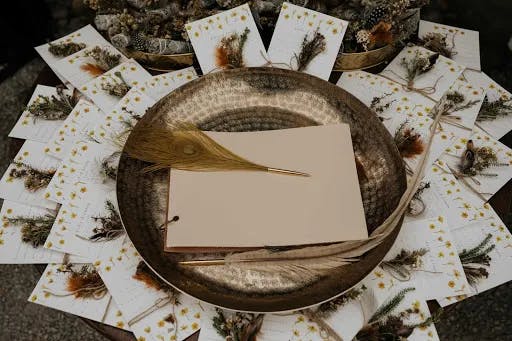 A flat lay of a gold decorative plate with a blank beige notebook and a gold pen resting on it. Surrounding the plate are botanical-themed cards, each adorned with dried flowers and feathers. The setup has a rustic and elegant appearance.