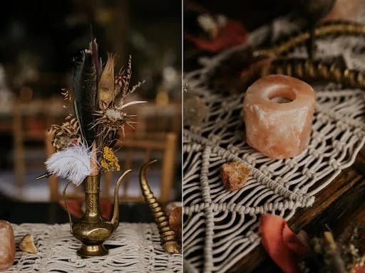 A rustic table setting with a brass teapot filled with dried flowers and feathers on the left. On the right, there's a pink Himalayan salt candle holder and a piece of raw amber on a woven table runner. The scene appears warm and earthy.