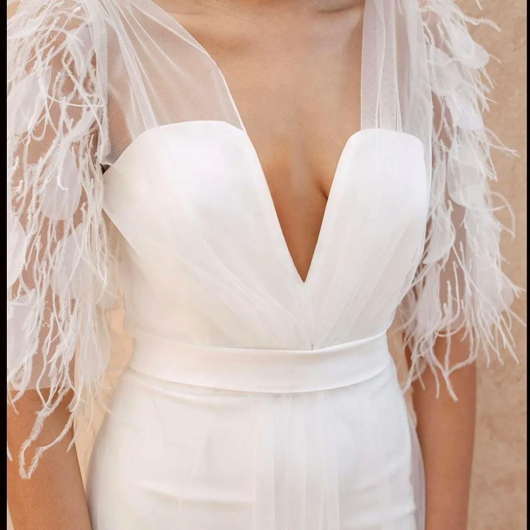 A person wearing a white wedding dress with a plunging neckline, sheer tulle overlay, and delicate feathered accents on the shoulders. The image captures the upper torso and the intricate details of the dress.