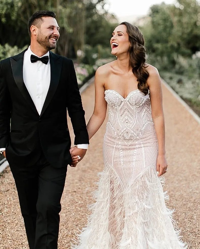 A joyful bride and groom walk hand in hand down a path outdoors. The bride wears a detailed, strapless white wedding gown, and the groom is in a black tuxedo with a bow tie. Both are smiling and looking at each other. The background is filled with greenery.
