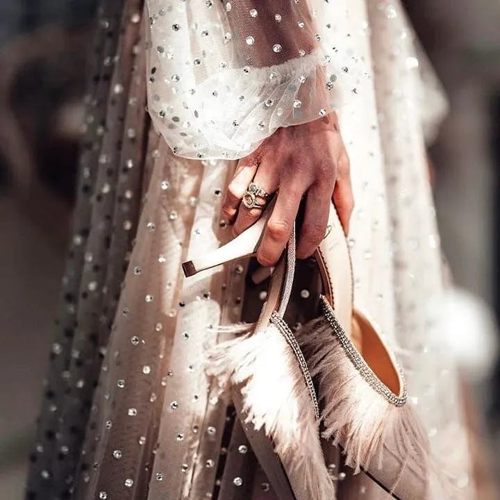 A person in a sparkling, sheer dress with polka dot patterns holds a pair of elegant high heels adorned with feathers and gems. The person wears a ring on the middle finger, while the sunlight casts a glowing effect on both the dress and the shoes.
